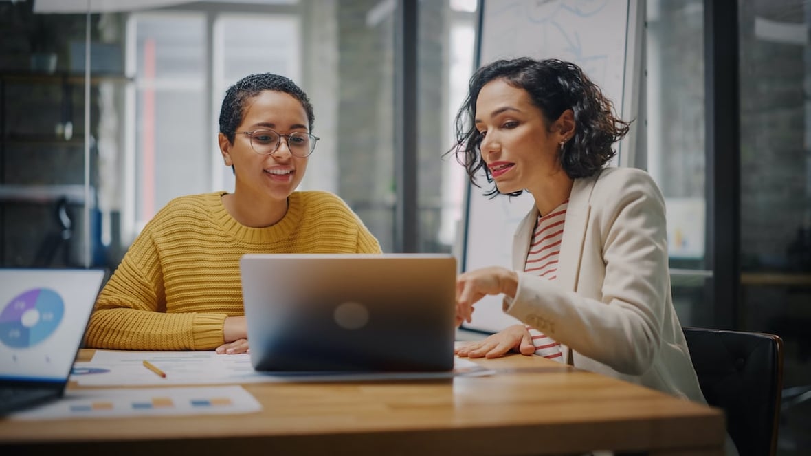 Two women sitting on a desk discussing something they see on their laptop's screen