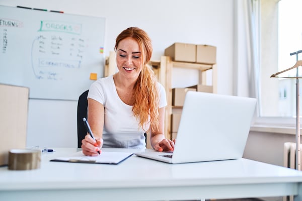 Caucasian woman sitting in an office while writing on a piece of paper with her laptop open