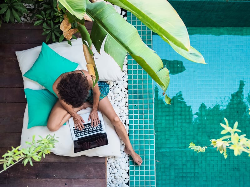 A person sitting beside a swimming pool and working remotely on a laptop