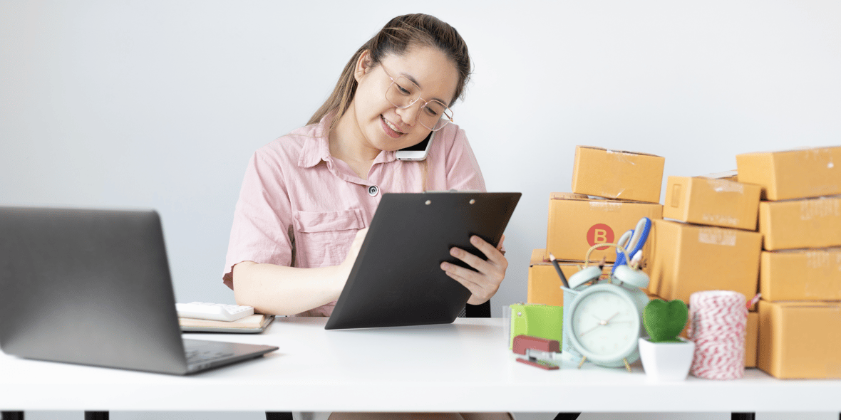 Woman looking at a checklist with a computer open to the side and boxes at the background