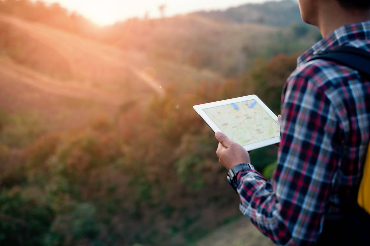 A young man standing on a hill overlooking a valley and holding a tablet with a navigational map on it illustrating the buyer's journey stages.