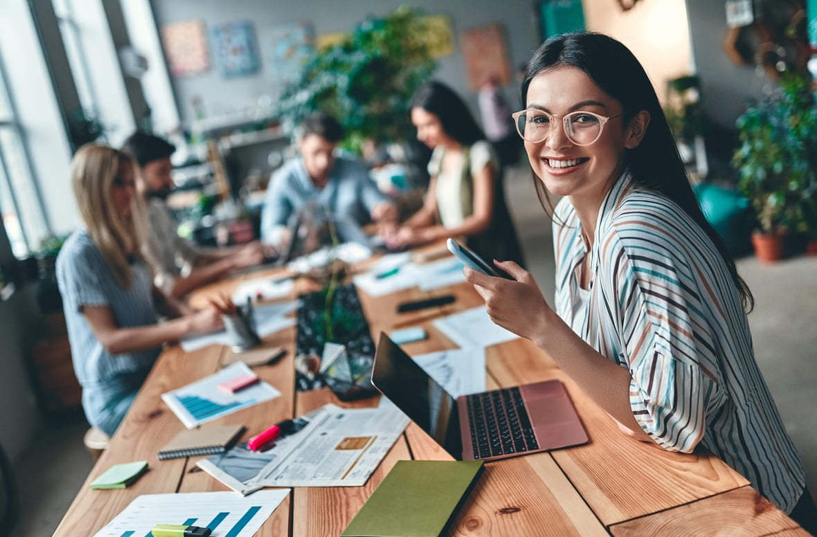 Woman smiling at the camera while sitting in a large table and working on her laptop