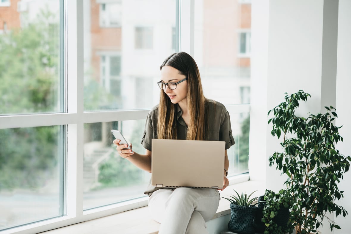 Caucasian woman looking at her phone screen while holding a laptop in her lap