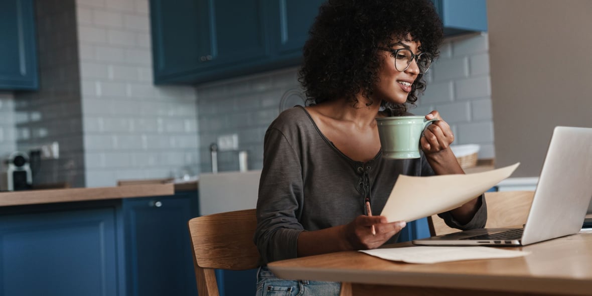 Woman sitting in her kitchen table looking at her laptop while drinking coffee